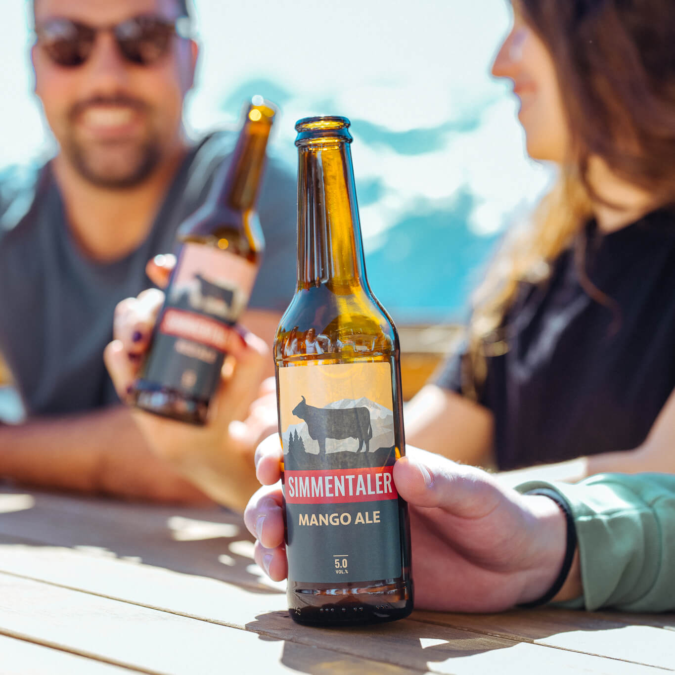 Lächelnde Menschen die an einem Holztisch im Freien ein Simmentaler Bier geniessen.